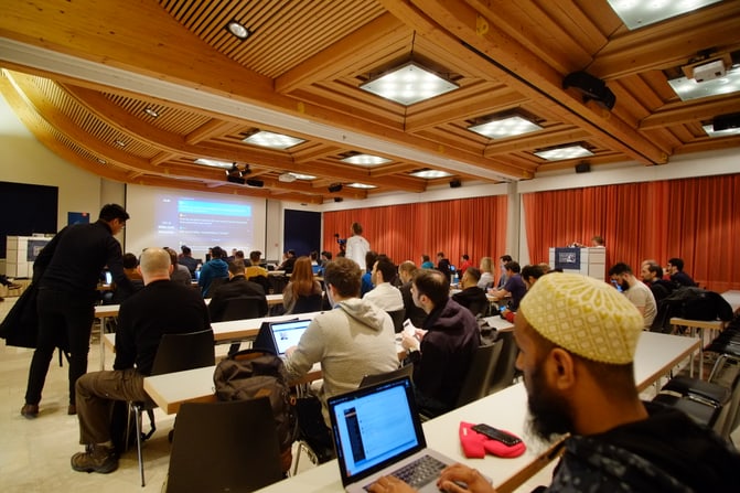 Mountain Camp Conference Room. A person with a muslim hat looking at the screen in the front and many other participants in a conference room. You can see a person with a video camera recording the front row
