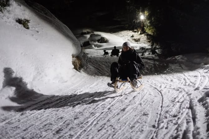 A cheerful-looking woman sledges down at night-lit slope. Another person sledding in the backround with a running dog next to it